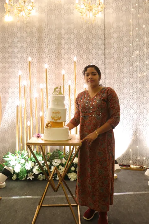 Athira Anoop, founder of Adhi's Bake House, beside a white and gold tiered wedding cake
