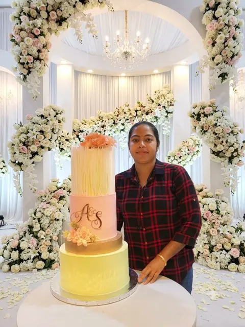 Athira Anoop, founder of Adhi's Bake House, with a four-tier wedding cake on stage