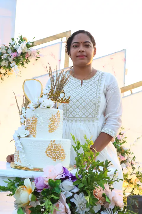 Athira Anoop, founder of Adhi's Bake House, with a white and gold wedding cake